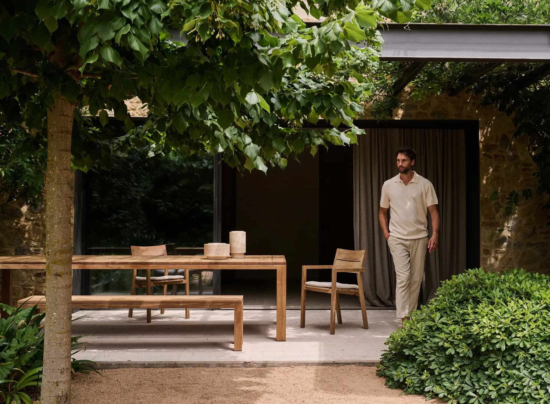 Tribù Kos teak dining table, bench and armchairs under a tree by a garden house